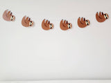 May include: A brown sloth garland with five sloths hanging from a white string. The sloths are facing to the right and have black eyes and noses.