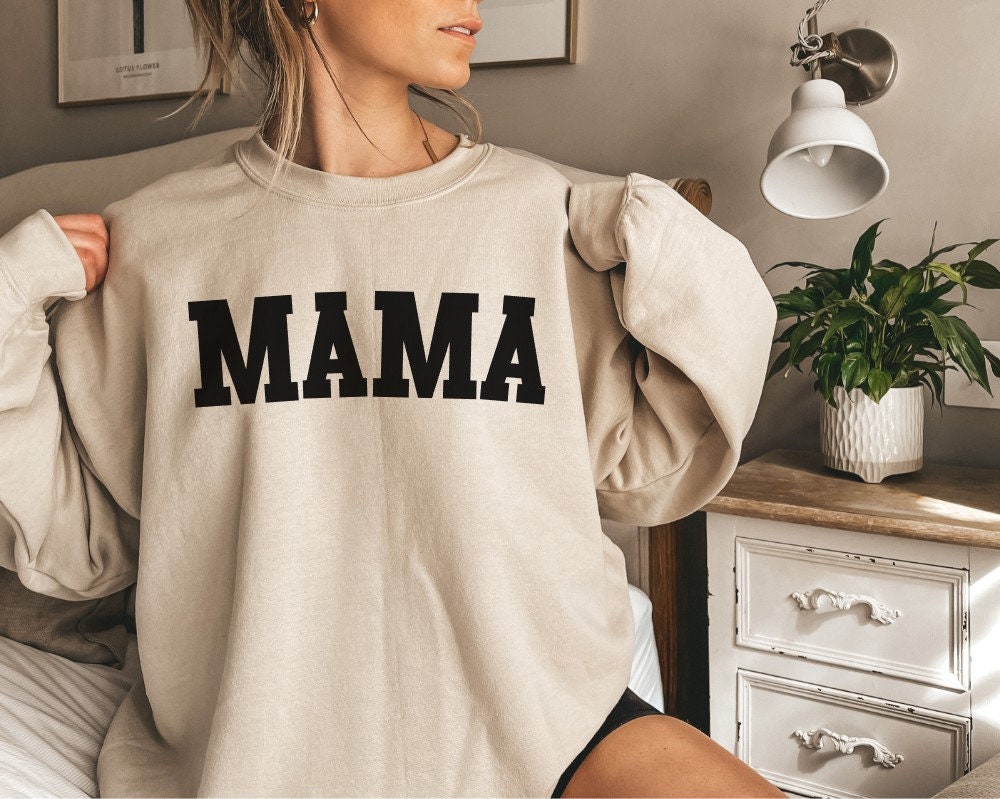 May include: A beige sweatshirt with the word 'MAMA' printed in black block letters on the front.