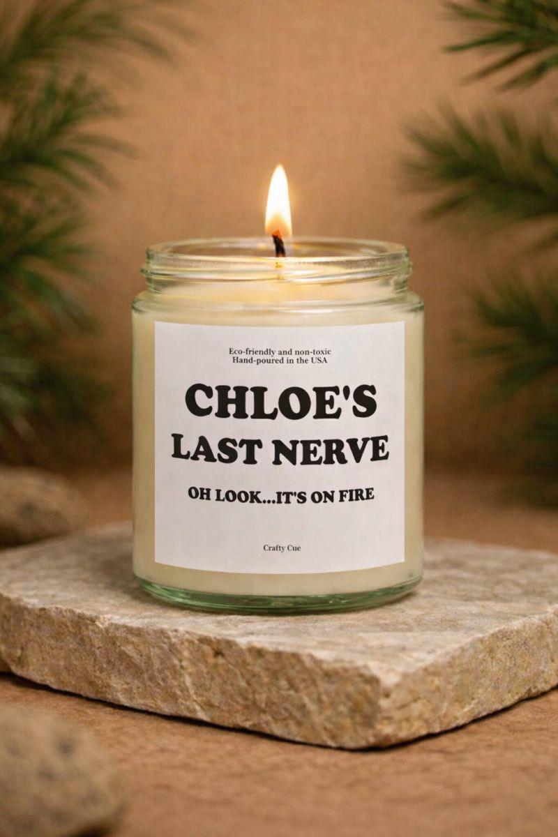 Personalized Last Nerve soy candle in a clear glass jar with a clean white label featuring bold black text that reads a custom name followed by “Last Nerve” and the phrase “Oh look… it’s on fire.” The candle is lit with a small flame and sits on a natural stone surface with soft greenery in the background. This funny scented candle makes a perfect gift for her or him, ideal for Christmas, birthdays, coworkers, friends, or anyone who loves sarcastic humor and modern home decor.