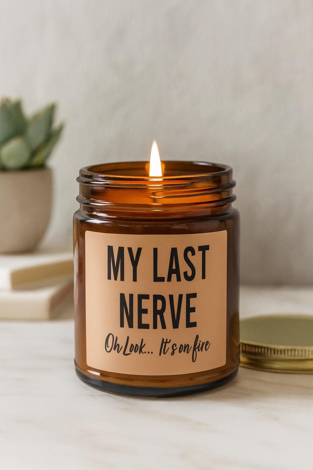 May include: A lit brown glass jar candle with a tan label. The label reads "MY LAST NERVE" above "Oh Look... It's on fire" in black text. A gold lid sits next to the candle. A small succulent plant is in the background.
