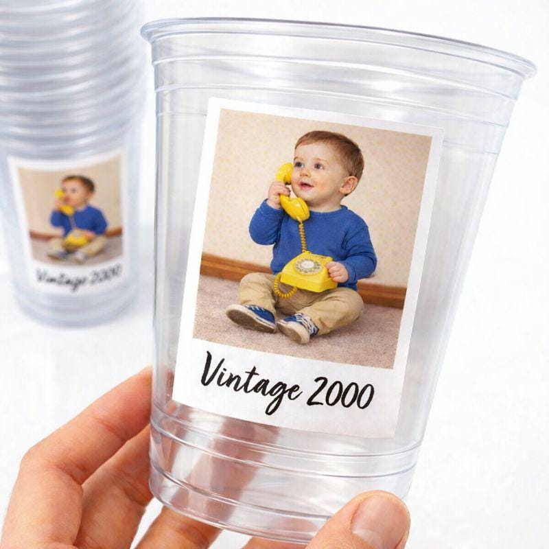May include: Clear plastic cups with a printed label featuring a baby holding a yellow rotary phone. The label reads "Vintage 2000". The baby is wearing a blue shirt and khaki pants. The cups are held by a hand.