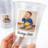 May include: Clear plastic cups with a printed label featuring a baby holding a yellow rotary phone. The label reads "Vintage 2000". The baby is wearing a blue shirt and khaki pants. The cups are held by a hand.