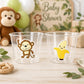 May include: Two clear plastic cups with cartoon stickers of a monkey and a banana. A plush monkey and a sign that says "Baby Shower" are in the background. Balloons and a wooden toy are also visible.