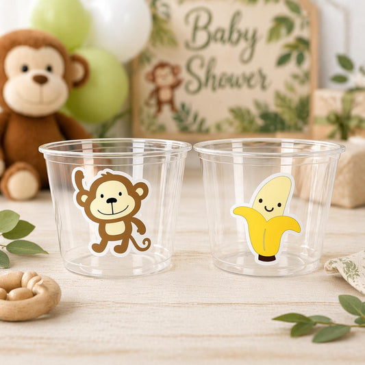 May include: Two clear plastic cups with cartoon stickers of a monkey and a banana. A plush monkey and a sign that says "Baby Shower" are in the background. Balloons and a wooden toy are also visible.