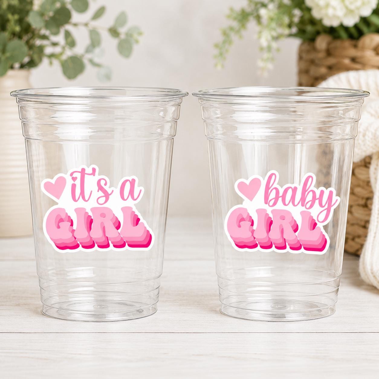 May include: Two clear plastic cups with pink text that reads "It's a GIRL" and "baby GIRL". The text is layered with a heart above the word "baby". The cups are for a baby shower or gender reveal party.