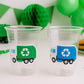 May include: Two clear plastic cups with recycling truck stickers, one green and one blue. A green toy garbage truck and green decorations are in the background. Chocolate chip cookies are on a plate. Party supplies.