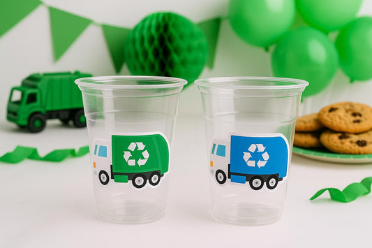 May include: Two clear plastic cups with recycling truck stickers, one green and one blue. A green toy garbage truck and green decorations are in the background. Chocolate chip cookies are on a plate. Party supplies.