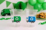 May include: Two clear plastic cups with recycling truck stickers, one green and one blue. A green toy garbage truck and green decorations are in the background. Chocolate chip cookies are on a plate. Party supplies.