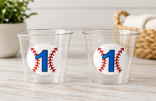 May include: Two clear plastic cups with a baseball-themed design. Each cup features a white circle with a blue number 1 and red baseball stitching. The cups are on a light-colored wooden surface.