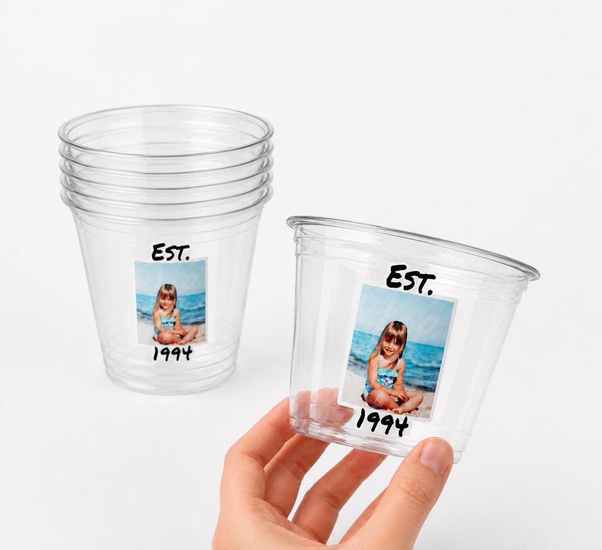 May include: Clear plastic cups with a photo of a young girl on the beach, the text "EST. 1994" is printed on the cups. One cup is stacked with others, and another is held by a hand.