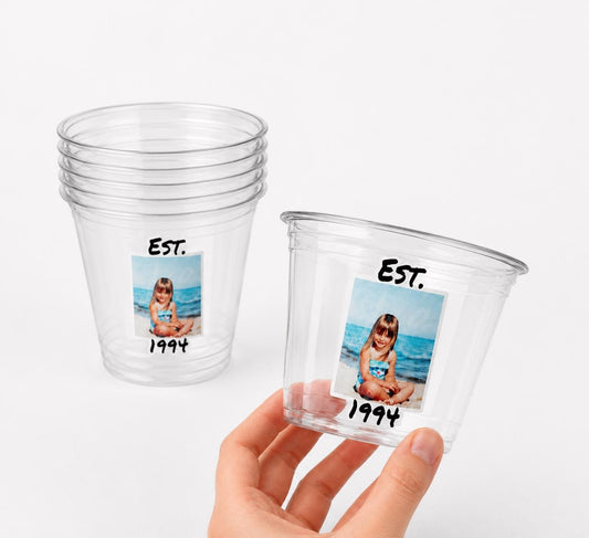 May include: Clear plastic cups with a photo of a young girl on the beach, the text "EST. 1994" is printed on the cups. One cup is stacked with others, and another is held by a hand.