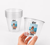 May include: Clear plastic cups with a photo of a young girl on the beach, the text "EST. 1994" is printed on the cups. One cup is stacked with others, and another is held by a hand.