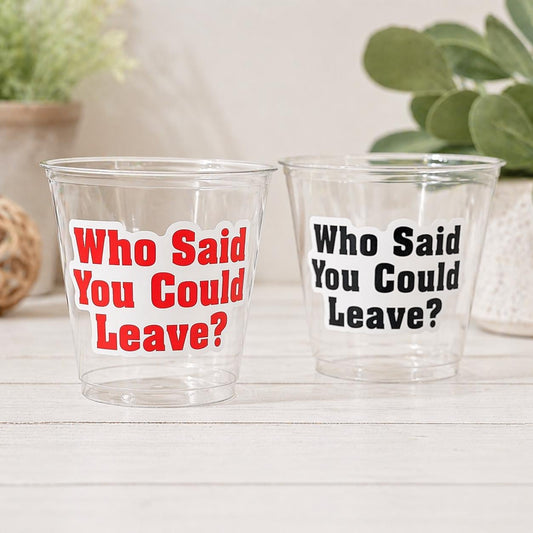 May include: Two clear plastic cups with the text "Who Said You Could Leave?" in red and black. The cups are on a white wooden surface, with greenery in the background.